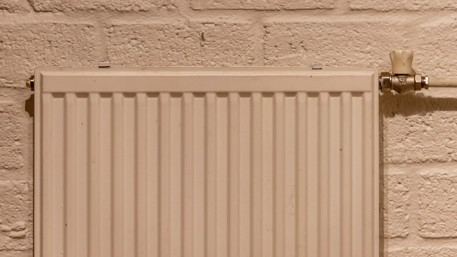 Weißer Plattenheizkörper mit Thermostat an einer hellen Backsteinwand.