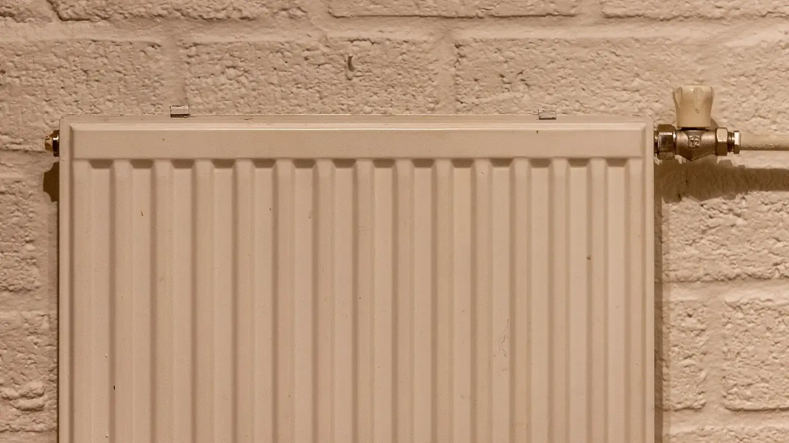 Weißer Plattenheizkörper mit Thermostat an einer hellen Backsteinwand.